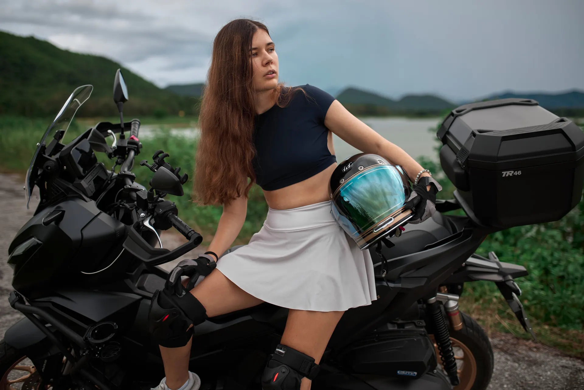 Nana holding a helmet beside a black motorcycle near a lake