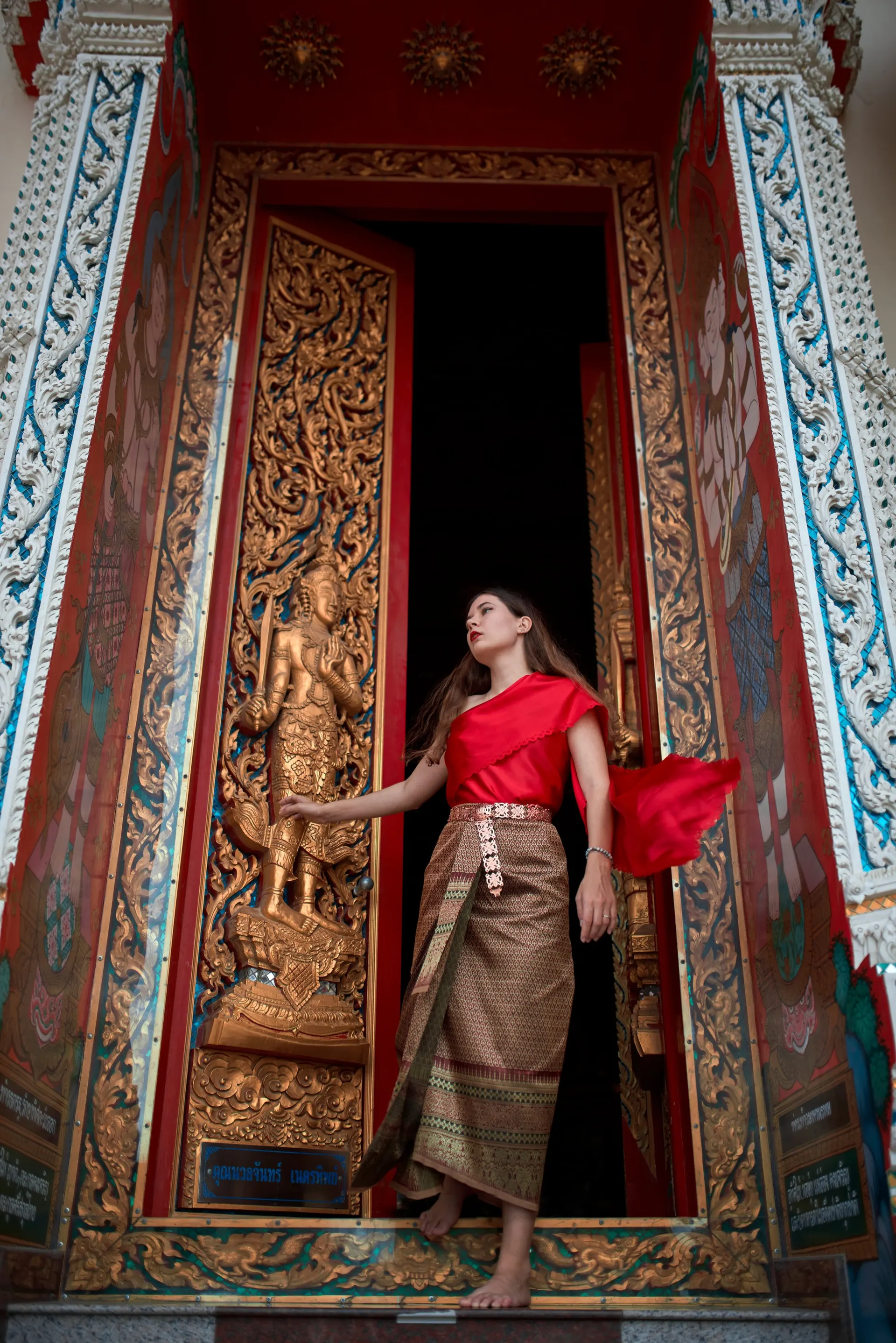 Nana in traditional Thai dress stepping out of an ornate gilded temple doorway
