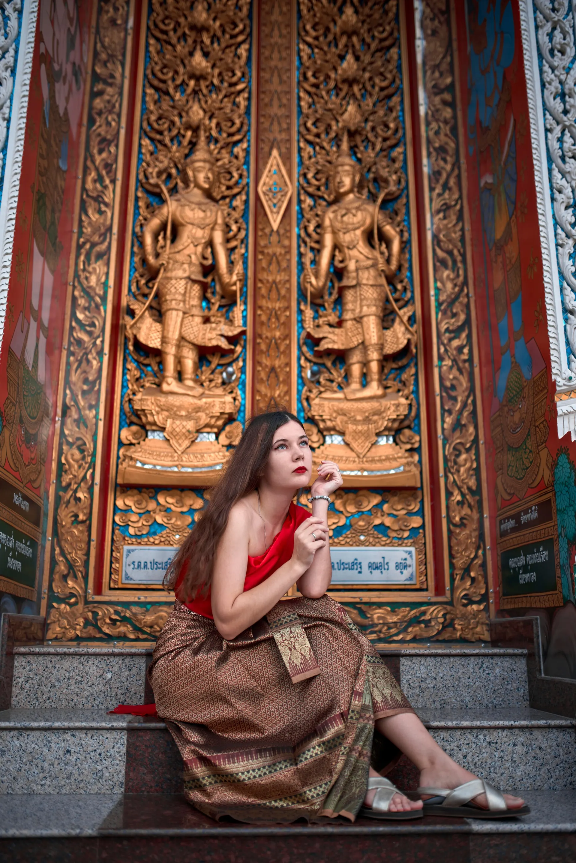 Nana in traditional Thai dress seated on temple steps below golden guardian figures